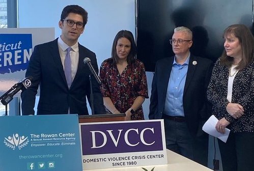 A man speaks into a microphone in front of a sign that says "Domestic Violence Crisis Center." Three others stand near, listening.